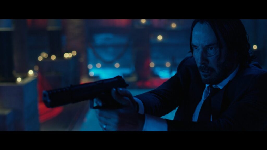 A screencap from one of the John Wick films, showing the titular character in a blue and red lit scene with yellow string lights holding up his gun, dressed all in black.