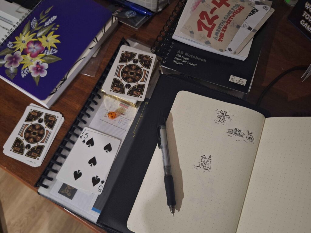 A photo of a very messy desk, with notebooks scattered around and a cameo of my small mental game 12-4 which you can get at reyesraine.itch.io (nudge nudge).

However the important thing is the deck of cards, with a pile for the cards I played for landmarks, and a discard pile, the d20, for my homebrew rule, and the notebook where I was drawing the map with a simple ballpoint pen.